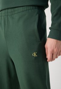 Mørkegrønne joggebukser laget av myk stoff med flat midje. Har en liten gull "cK"-logo på venstre side og sidelommer.