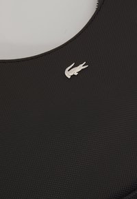 Gros plan sur un tissu noir texturé avec un petit emblème crocodile argenté près de l'encolure arrondie.