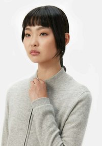 Ljusgrå, ribbad stickad tröja med hög krage och långa ärmar. Modellen håller försiktigt i kragen, vilket visar den mjuka texturen.