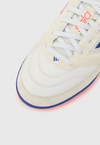 Λευκό αθλητικό παπούτσι με επάνω μέρος από σουέντ και διχτυωτό υλικό, με μοβ λεπτομέρειες, υφασμένα επικαλύμματα και λευκά κορδόνια. Η εξωτερική σόλα έχει εμφανή αντίθεση χρώματος.