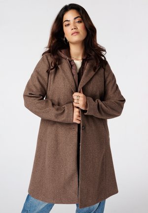 Jeune femme aux longs cheveux foncés portant un manteau en laine marron par-dessus un sweat à capuche marron et un jean bleu, debout devant un fond clair uni.