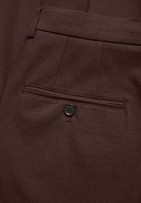 Pantalones marrones con una textura suave, un bolsillo trasero con un botón negro y detalles de pliegue sutiles en la tela.