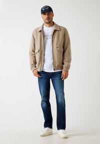 Beige wollen blend ritsjack met twee, voorzakken, witte T-shirt met logo, blauwe spijkerbroek en witte sneakers. Donkere pet met logo.