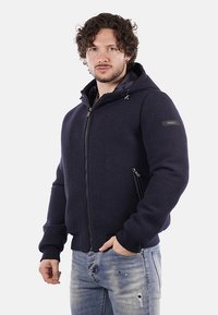 Giacca a cappuccio blu navy con chiusura a zip, dotata di tasche laterali e polsini a coste. Realizzata in materiale testurizzato con finitura opaca.