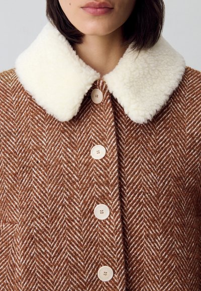 Abrigo marrón con patrón de espiga, con un cuello de pelo sintético color crema y botones claros. La tela tiene una textura suave y un diseño estructurado.