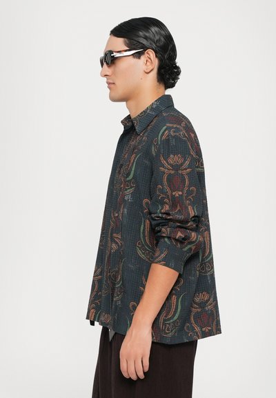 Button-up shirt with a dark textured fabric featuring floral and abstract patterns in brown and green; model wearing sunglasses.