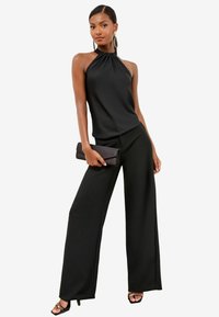Melna halter top, kas apvienots ar platiem melniem biksēm, kas izskatās eleganti un gludi. Tur rokās melnu clutch somiņu.