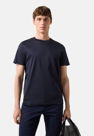 Ung mand iført en ensfarvet mørk marineblå t-shirt og bukser, holder en sort lædertaske i højre hånd mod en lys baggrund.