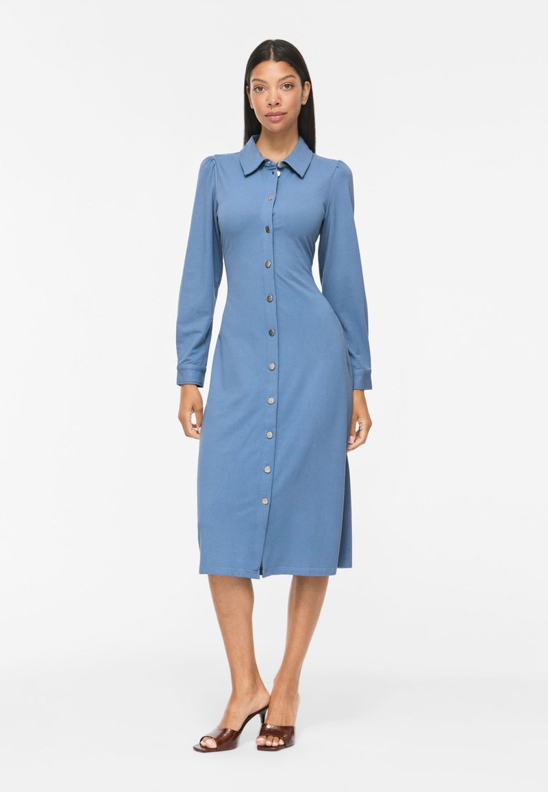 Femme portant une robe midi bleue à manches longues, boutonnée sur le devant avec un col, associée à des sandales marron ouvertes à talons, debout sur un fond blanc.