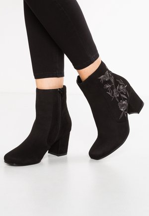 Sorte ruskind ankle støvler med blokhæle, sidelukning med lynlås og blomstret broderi på ydersiden, båret med sorte tætsiddende bukser.