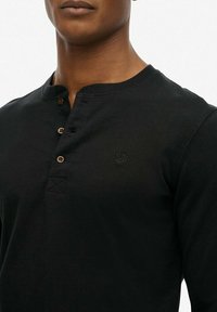 Zwarte lange mouwen henley-shirt gemaakt van zachte katoen. Heeft een ronde halslijn, drie bruine knopen en een klein logo op de borst.