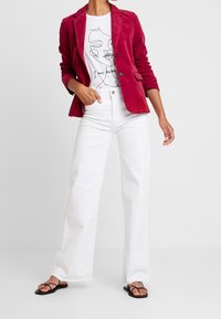 Nainen, joka on pukeutunut punaiseen samettitakkiin, valkoiseen graafiseen t-paitaan, jossa lukee "Olen oma itseni", valkoisiin leveälahkeisiin farkkuihin ja mustiin nauhakorkokenkiin, seisoo käsi taskussa.