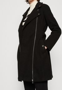 Svart kappa med asymmetrisk dragkedja, slagkrage och sidofickor. Tillverkad av strukturerad tyg, med silverfärgade detaljer.