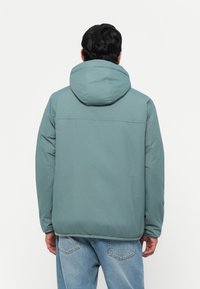 Lys-teal pufferjakke med hette, med en jevn tekstur og elastiske mansjetter. Baksiden viser en horisontal søm på tvers av midten.
