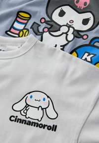 Biała bawełniana bluza z kapturem z motywem kreskówkowego królika z niebieskimi oczami i różowymi polikami, podpisana "Cinnamoroll", na jasnoszarym tle.