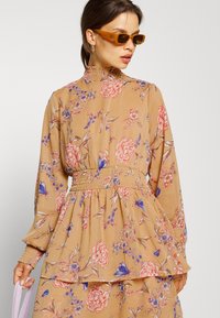 Vestido de manga longa e gola alta em tecido cor de areia, com padrões florais em rosa, vermelho e azul. Cintura franzida e detalhe de bainha em camadas.