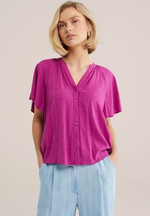 Ženska s kratkimi blond lasmi nosi svetlo magenta srajco z gumbi in krilci na rokavih ter svetlo modre hlače z visokim pasom, roke v žepih.