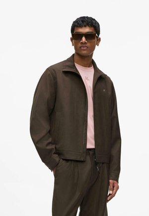 Hombre con chaqueta marrón oscura con cremallera y pantalones a juego, camisa rosa claro y gafas de sol oscuras y grandes, de pie frente a fondo blanco.