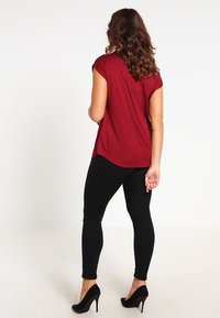 Kvinna med lockigt hår som bär en röd kortärmad topp, svarta leggings och svarta högklackade skor, stående med ryggen mot kameran.