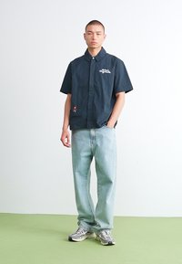 Kortermet svart skjorte med diskret logo-broderi, sammen med lyseblå denimbukser og grå joggesko, stående på et grønt gulv.