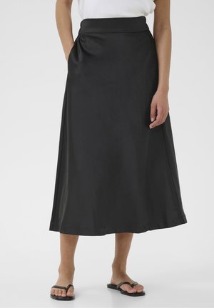 Kvinde iført sort midinederdel med lommer, hvid top og sorte flade sandaler, stående foran en ensfarvet hvid baggrund.