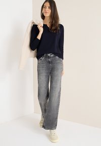 Meriino-pitkähihaisessa paidassa on napit, yhdistettynä väljähihaisiin, haalistuneisiin harmaisiin farkkuihin ja beigeihin tennareihin, seisoo neutraalin taustan edessä.