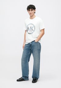 Camiseta de algodón blanca con un logotipo gráfico redondo en gris y azul. Combinada con unos pantalones vaqueros de mezclilla de corte holgado y color azul claro, y zapatillas negras.