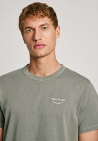 Aliejinė žalia trumpomis rankovėmis marškinėliai iš medvilnės; turi apvalią kaklo iškirptę ir mažą baltą "Pepe Jeans" logotipą ant kairiojo krūtinės.