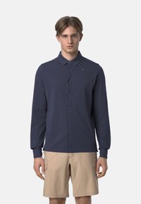 Camicia a maniche lunghe blu navy con colletto a punta, caratterizzata da un logo discreto sul petto e un tessuto liscio e strutturato. Indossata con pantaloni corti beige.