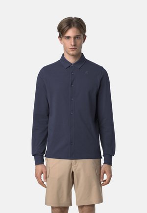 Camicia a maniche lunghe blu navy con colletto a punta, caratterizzata da un logo discreto sul petto e un tessuto liscio e strutturato. Indossata con pantaloni corti beige.