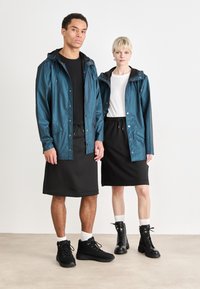 Vanntett teal-jakke med hette, svart sweat-skjørt og svarte støvler; modeller viser en uformell stil med kontrasterende farger og teksturer.