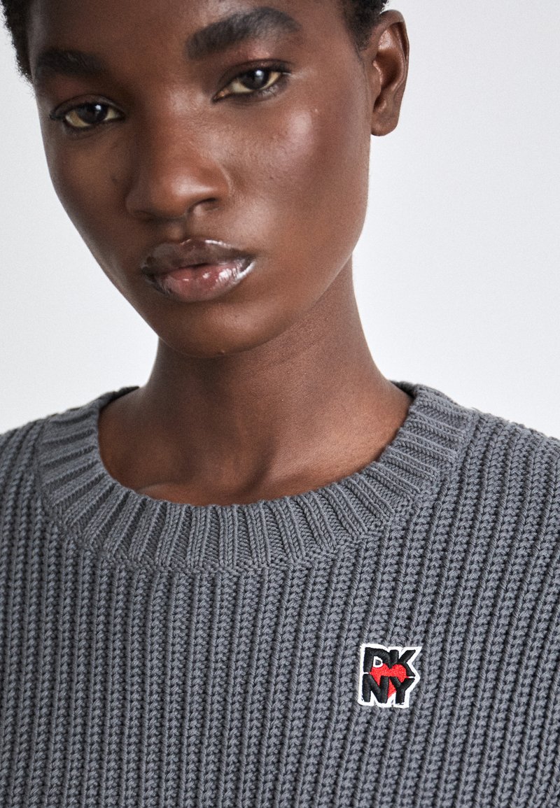 Grauer gerippter Pullover mit rundem Halsauschnitt. Mit einem rot-schwarzen gestickten Logo auf der Brust. Glatte Textur und gestricktes Design.