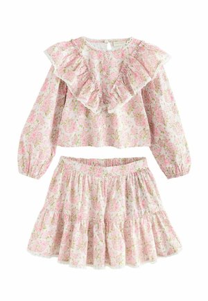 Mladé dievča s tmavými vlasmi v ružových kvetinových šatách s volánikmi na hrudi a sukni, stojace pred hladkým béžovým pozadím.