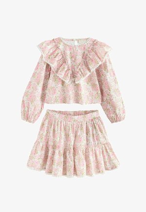 Mladé dievča s tmavými vlasmi v ružových kvetinových šatách s volánikmi na hrudi a sukni, stojace pred hladkým béžovým pozadím.