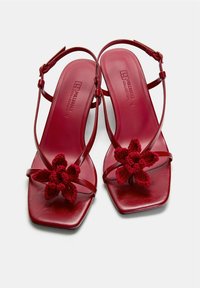 Sandales rouges avec un design à bout carré, dotées de fines lanières et d'une jolie fleur en crochet sur le devant. Texture en cuir lisse.