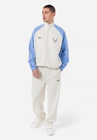 Balts zip-up treniņjaka ar gaiši ziliem piedurknēm un atbilstošām biksēm. Iekļauj logo izšuvumu un elastīgus aproces, kas nodrošina komfortu.