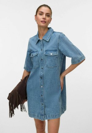 Naine kannab lühikeste varrukatega, nööbitud teksakleiti rinnataskutega, hoides ühes käes pruuni tutidega seemisnahast kotti.