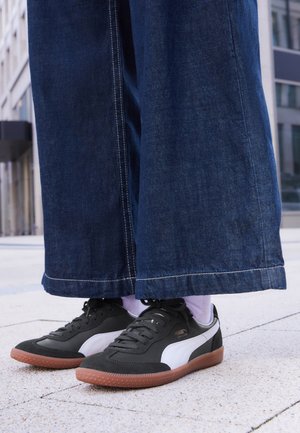 Person iført mørkeblå jeans med vide bukseben, hvide sokker og sorte Puma-sneakers med hvide striber og gummisåler, der står på fortovet.