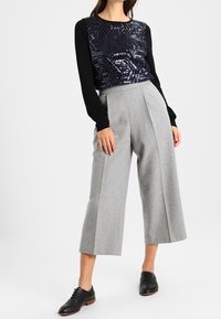 Femme portant un haut noir à manches longues avec un motif géométrique en sequins bleu marine, un pantalon large cropped gris clair, et des chaussures noires à lacets.