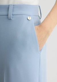 Helle blaue Hose aus glattem Stoff. Verfügt über eine Seitentasche und einen kleinen silbernen Akzentknopf in der Nähe des Bundes.