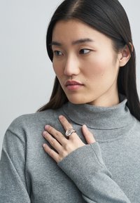 Srebrn prstan z velikim prozornim dragim kamnom, nošen na roki, ki počiva na sivem puloverju z visokim ovratnikom in rebrastimi manšetami. Ton kože je svetel do srednji.