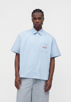 Joven con el pelo trenzado que lleva una camisa de manga corta azul claro con botones y texto rojo en el bolsillo del pecho y pantalones vaqueros claros.
