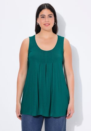 Frau mit dunklen Haaren trägt ärmelloses, türkisfarbenes, plissiertes Oberteil und blaue Jeans, steht vor einem hellen Hintergrund und schaut leicht lächelnd nach vorn.