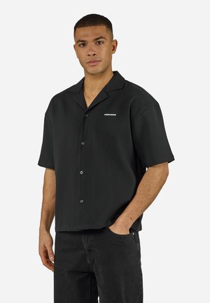 Jeune homme portant une chemise noire à manches courtes avec de fines rayures verticales et un pantalon noir, debout avec une main dans la poche.