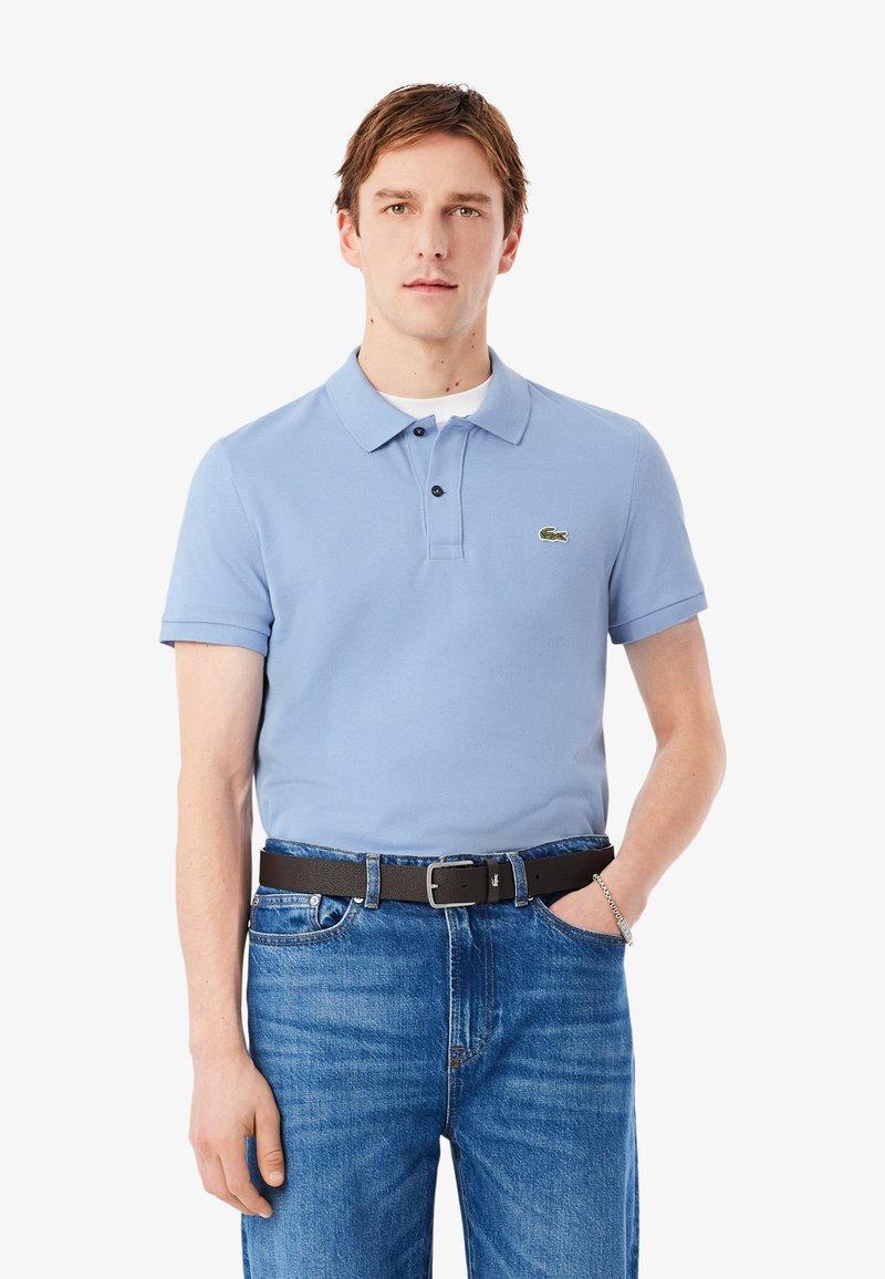Hellblaues Poloshirt mit Kragen, zwei schwarzen Knöpfen und einem grünen, bestickten Logo, kombiniert mit blauen Jeans und einem schwarzen Gürtel.