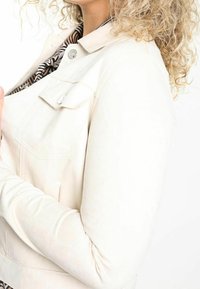 Cremefarvet jakke med en tætsiddende design, der fremhæver en krave, brystlomme og teksturerede ærmer. Stof virker glat og struktureret.