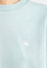 Sudadera de punto azul claro con pequeño logo blanco bordado en el pecho, mostrando la parte superior del torso y el escote.