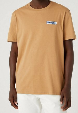 Person wearing a tan Wrangler logo t-shirt with short sleeves and white pants, standing against a light background.