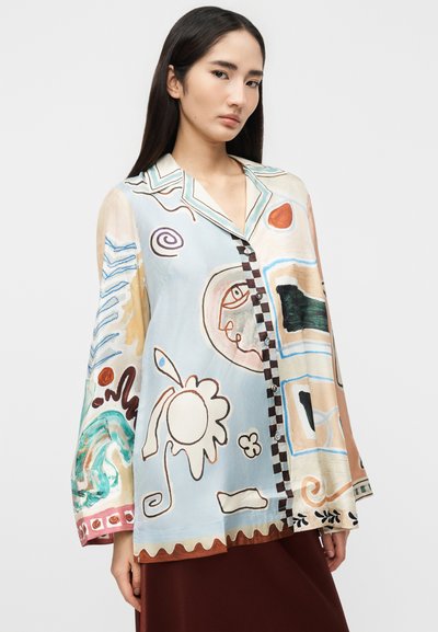 Νεαρή γυναίκα που φοράει μια χαλαρή, πολύχρωμη πουκάμισα με αφηρημένο μοτίβο και μακριά μανίκια, στέκεται μπροστά σε ένα απλό λευκό φόντο.