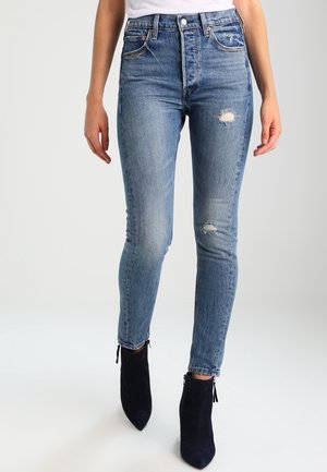 Kvinde iført lyseblå slidte skinny jeans og sorte ankelstøvler, stående foran en ensfarvet lysegrå baggrund.